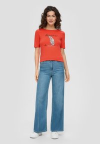 Czerwona koszulka z krótkim rękawem z grafiką i napisem, w zestawieniu z szerokimi, jasnoniebieskimi dżinsami i białymi trampkami.