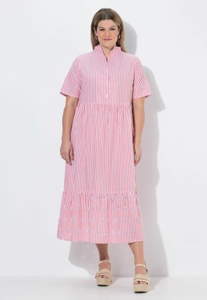 Kobieta stojąca w różowo-białej sukience midi w paski z krótkimi rękawami i haftowanymi detalami, w połączeniu z beżowymi sandałami na platformie.
