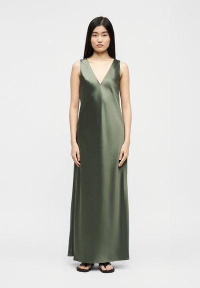 Γυναίκα με μακριά μαύρα μαλλιά που φοράει αμάνικο, μάξι φόρεμα από σατέν σε ελιά πράσινο χρώμα και μαύρα σανδάλια, στέκεται μπροστά σε έναν απλό άσπρο φόντο.