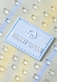 Ljusblå "Johnny Urban" etikett med glasögonlogotyp på texturerad tyg med små transparenta pärlor och pastellrandigt mönster.