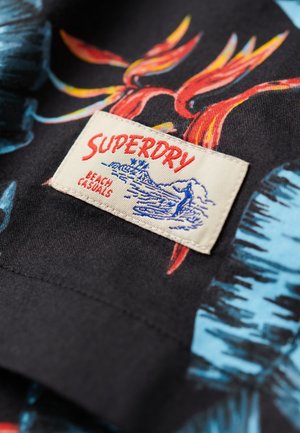 Tessuto nero con vivaci motivi tropicali blu e rossi; un'etichetta color crema riporta la scritta "SUPERDRY BEACH CASUALS" con un'illustrazione del logo.