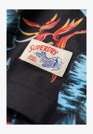 Tela negra con vibrantes patrones tropicales azules y rojos; una etiqueta de color crema dice "SUPERDRY BEACH CASUALS" con una ilustración del logo.
