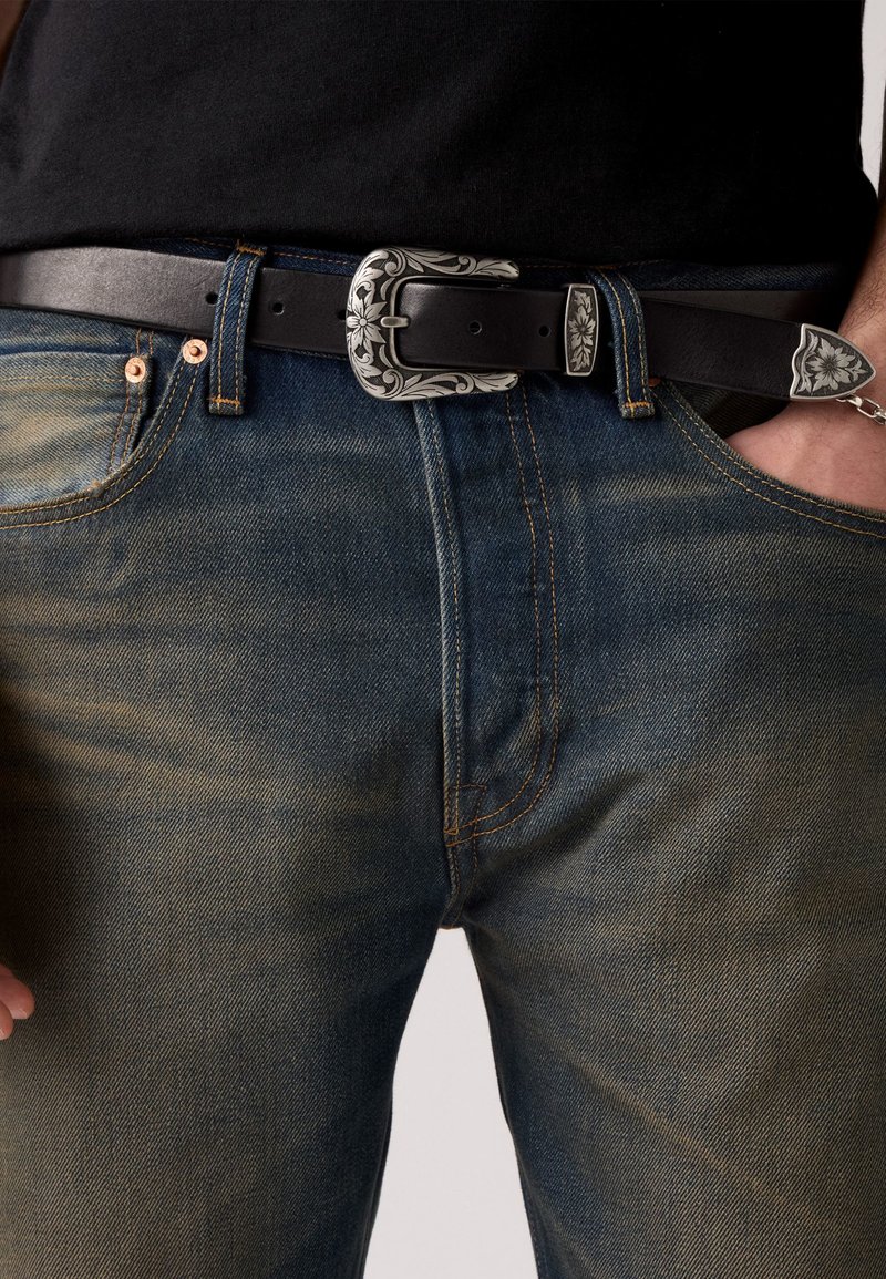 Ceinture en cuir noir avec une boucle en argent ornée, portée sur un jean en denim bleu délavé avec des rivets en cuivre et des détails de couture jaunes.