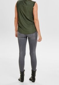 Olijfgroene mouwloze blouse met een gegolfde halslijn, gecombineerd met strakke grijze leggings en zwarte enkellaarsjes.