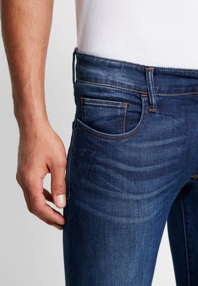 Mörkblå denimjeans med smal passform, med en subtil textur, orange sömmar och en liten framficka. Handen placerad vid midjan.