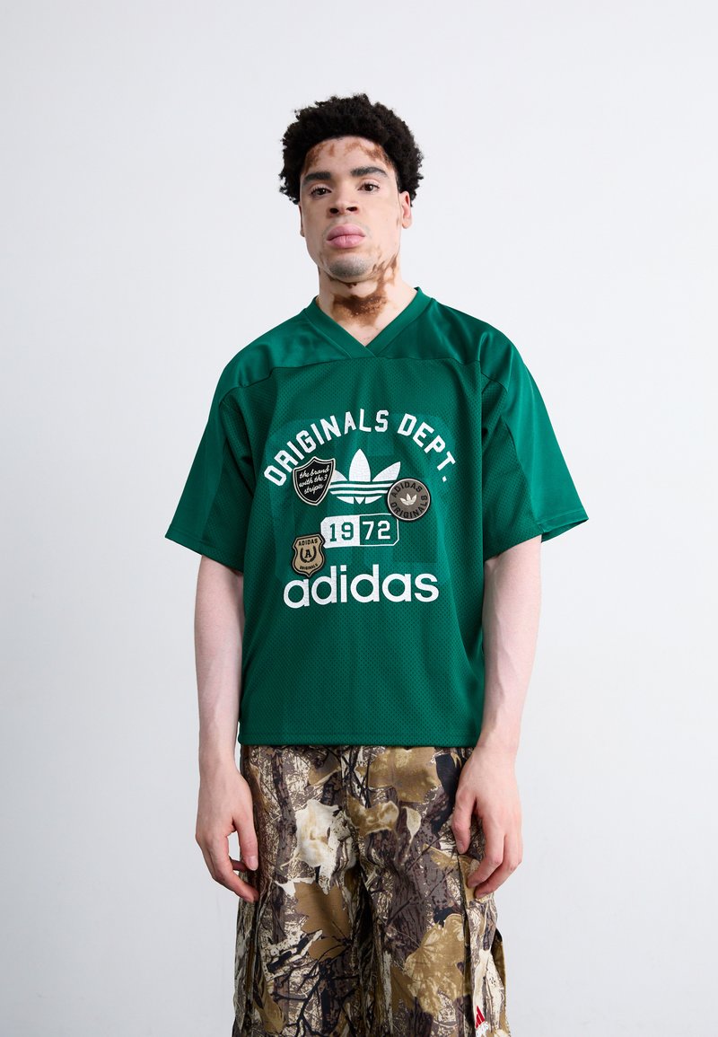 Zöld hálós mez, rajta "ORIGINALS DEPT" és "adidas" felirat, patchekkel és V-vágású fazonnal. Kamuflázs mintás sorttal párosítva.