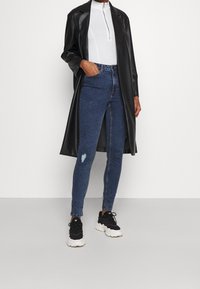 Černý kabát z umělé kůže, bílý top s zipem u krku, opotřebené modré skinny džíny a černé tenisky s bílou podrážkou.