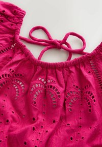 Tissu ajouré rose vif avec des motifs floraux brodés et un cordon en tissu noué au niveau de l'encolure froncée d'un vêtement.