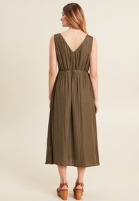 Olivgrünes, ärmelloses Kleid mit V-Ausschnitt, elastischem Taillenzug und fließendem Design, kombiniert mit braunen Keilsandaletten.