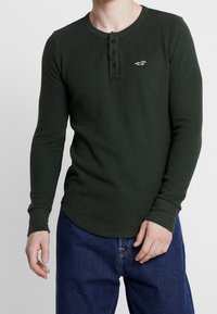 Camiseta henley de manga larga verde oscuro con un tejido texturado, tres botones negros y un pequeño logo blanco en el lado izquierdo del pecho. Combinada con jeans azules.