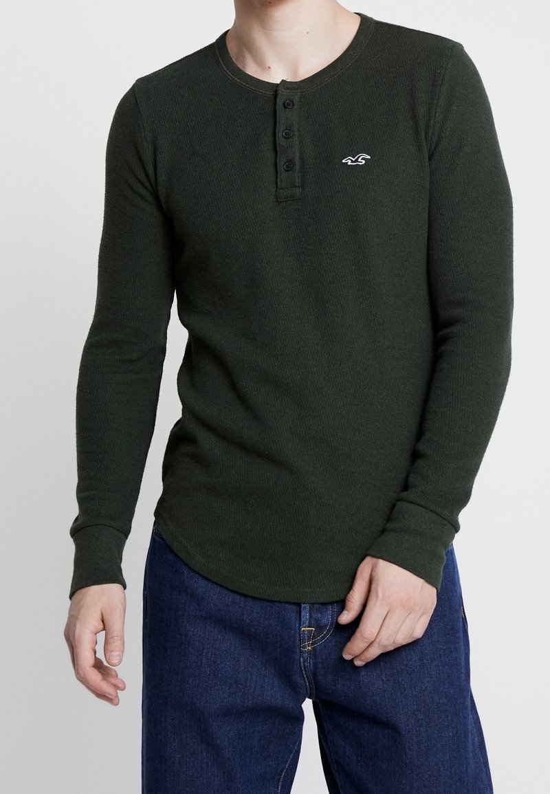 Camiseta henley de manga larga verde oscuro con un tejido texturado, tres botones negros y un pequeño logo blanco en el lado izquierdo del pecho. Combinada con jeans azules.