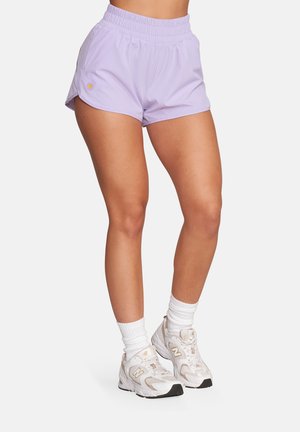 Kobieta ubrana w jasnofioletowe sportowe szorty, białe skarpetki za kostkę i białe buty New Balance, stojąca na tle jednolitego tła.