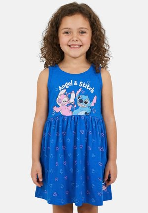 Fille souriante aux cheveux bouclés portant une robe bleue sans manches avec les personnages Angel et Stitch et un motif de cœur.