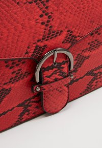 Punamusta käärmekuvioitu keinonahkalaukku, jossa kiiltävä musta solkikoriste ja sileä pinta. Kompakti suorakaiteen muoto.