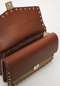 Bruine leren handtas met een gouden sluiting, gestepte accenten en een geweven textuur aan de onderkant. Het interieur heeft een gemusterde voering.