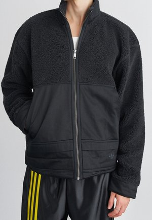 Person wearing a black fleece jacket with a front zipper and pockets, paired with black pants featuring yellow side stripes.