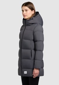 Pufová bunda v tmavošedej farbe s kapucňou, s prešívaným dizajnom a predným zipsom. Obsahuje bočné vrecká a nášivku s logom na leme.