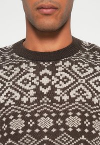 Dunkelbrauner und cremefarbener Pullover mit geometrischem Muster. Verfügt über einen gerippten Halsausschnitt und ein strukturiertes Strickdesign, das sich über den Stoff erstreckt.