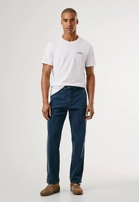 Fehér pamut póló kis logóval, sötétkék farmernadrággal párosítva. A modell barna cipőt visel, semleges háttér előtt áll.