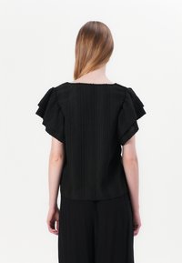 Musthave mustre levige varrukatega must topp, laineteta tekstuuriga kangas ja ruudukujuline lõige, koos mustade pükstega.