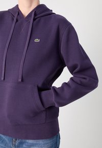 Violetinė Gobtuvo megztinis, pagamintas iš storos medžiagos, su kengūros kišene, spiralinėmis rankovėmis ir mažuoju žaliu logotipu ant krūtinės.