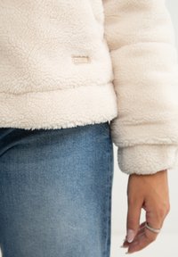 Veste en polaire beige clair avec une surface texturée, comportant une petite étiquette de marque. Assortie à un jean en denim bleu. Vue rapprochée de la manche.