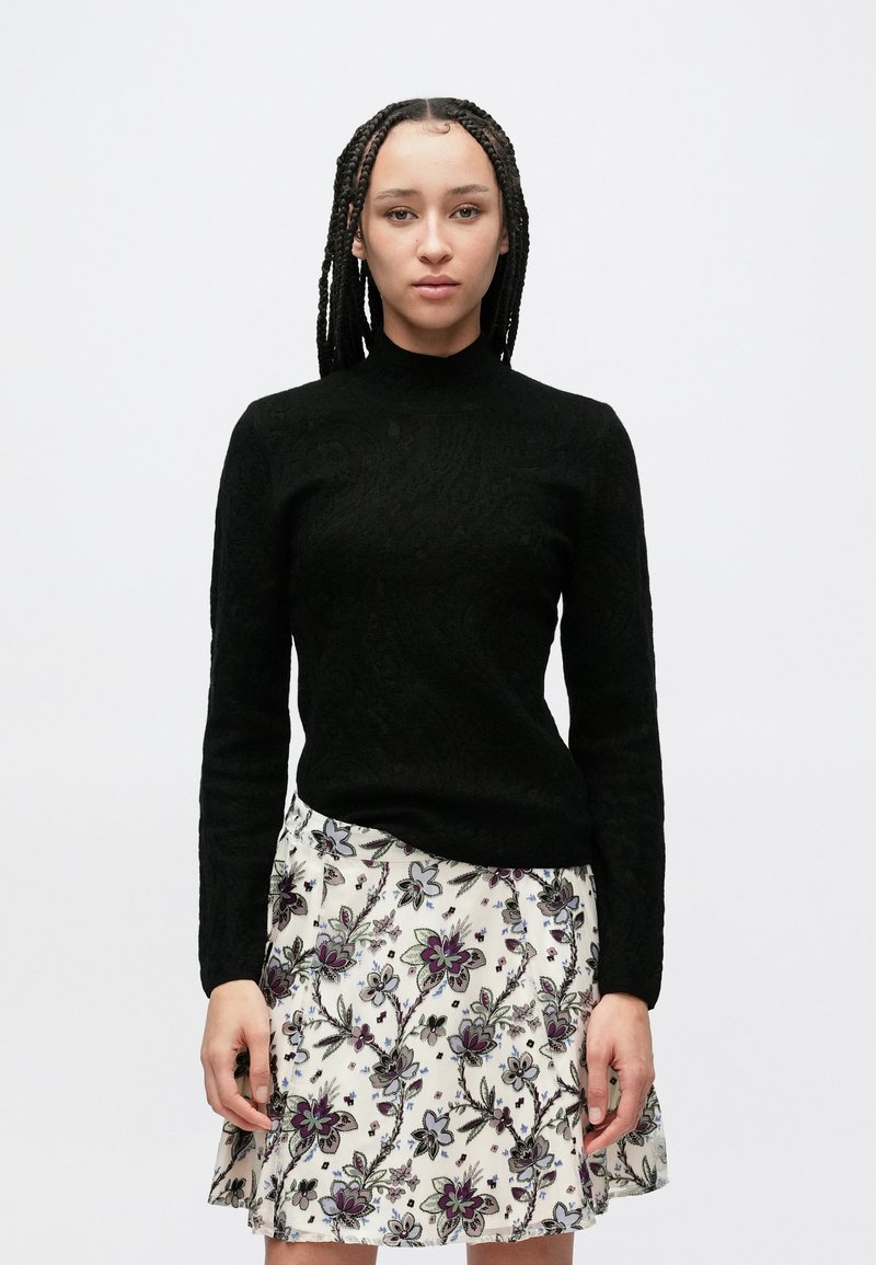 Μαύρο πουλόβερ με μακριά μανίκια και υφασμάτινη λεπτομέρεια, σε συνδυασμό με φούστα με λουλουδένιο σχέδιο που περιλαμβάνει μοβ και πράσινες αποχρώσεις σε ανοιχτό φόντο.