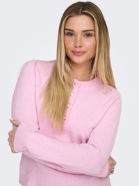 Mlada ženska z dolgimi blond lasmi, oblečena v roza pulover s perličnimi gumbi, zloženih rok, rahlo se smehlja na svetli podlagi.