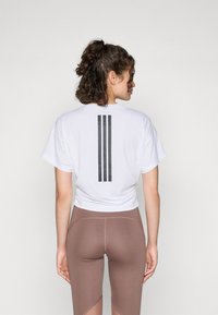T-shirt bianco cropped con logo a tre strisce nere sul retro, abbinato a leggings marroni a vita alta con un design color-block.