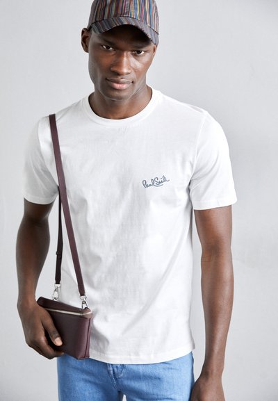 Valge puuvillane T-särk, millel on tikandiga logo, koos siniste denimitega. Mudel hoiab marooni värvi nahkot koos pika rihmaga.