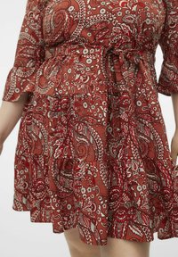 Robe rouge à motif cachemire avec une jupe évasée, une taille élastique et un détail à nouer. Présente des manches en cloche et des accents floraux tout au long.