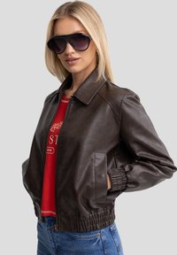 Giubbotto bomber in pelle marrone con colletto, polsini elasticizzati e chiusura con zip, abbinato a occhiali da sole oversize scuri e una camicia rossa.