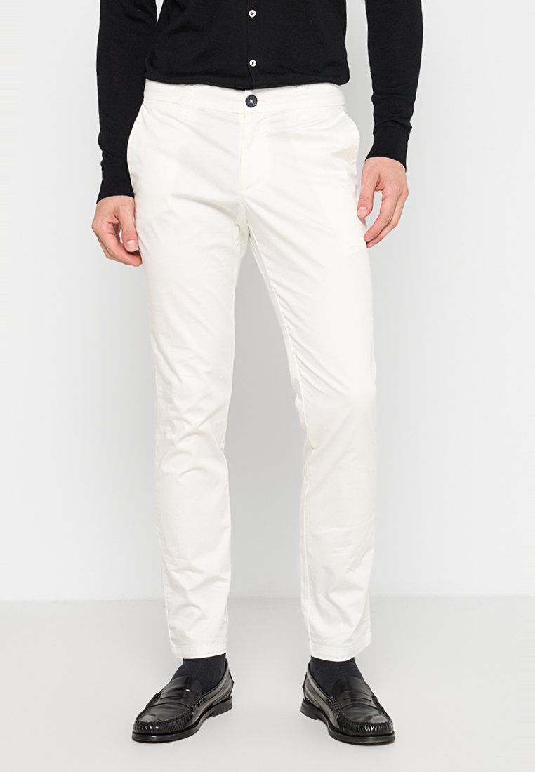 Pantaloni chinos bianchi in cotone con vestibilità slim, dotati di tasche laterali, chiusura con bottone e una texture liscia. Indossati con mocassini neri e calzini.