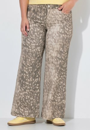 Personne portant un pantalon à jambes larges beige et marron à motifs, une main dans la poche et des baskets jaunes sur un fond uni.