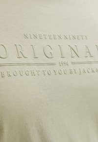 Μπεζ ύφασμα με ανάγλυφο κείμενο που γράφει "NINETEEN NINETY ORIGINAL 1990 BROUGHT TO YOU BY JACK&amp;" σε μινιμαλιστικό στυλ.