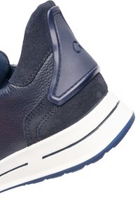 Detailní záběr paty sportovní obuvi v námořnické modři, zobrazující strukturovanou kůži, semišové panely, plastovou oporu paty a bílou gumovou podrážku s modrým vzorkem.