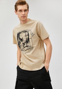 Camiseta de algodón color beige con un estampado gráfico de un cráneo humano y marcas de medición, que cuenta con un cuello redondo y mangas cortas.