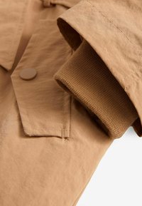 Gros plan d'une manche de veste beige avec une poche à rabat boutonnée et une bord-côte en maille marron qui dépasse de l'intérieur de la manche.