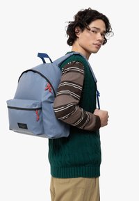 Un sac à dos bleu clair avec des accents noirs, une poche avant et des zips rouges. Le modèle porte un gilet en tricot vert sur une chemise à rayures marron.