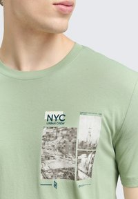 Artimas plano žmogaus, dėvinčio šviesiai žalią marškinėlį su juodai baltomis Niujorko miesto peizažo iliustracijomis ir užrašu "NYC Urban Crew."