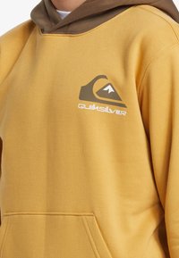 Gul hoodie med bruna detaljer, har en frampocket och en Quiksilver-logotyp tryckt i vitt och brunt. Mjuk tygkvalitet.