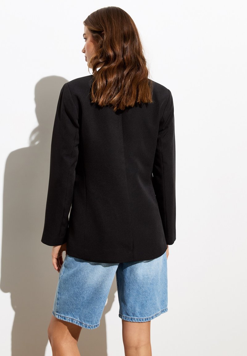 Blazer noir ajusté avec des manches longues, associé à un short en denim clair. Texture lisse, design cintré et détails minimalistes.