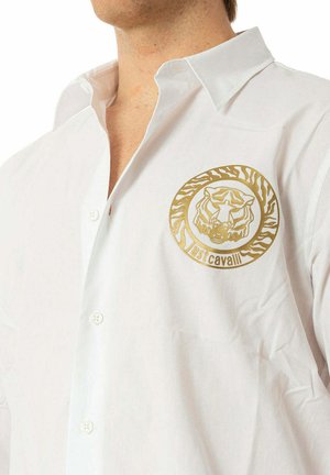Mężczyzna w białej koszuli na guziki z złotym logo głowy tygrysa i napisem „Just Cavalli” na piersi.