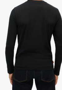 Person sedd bakifrån iförd en svart långärmad skjorta och mörkblå jeans med synlig orange sömnad på fickorna.