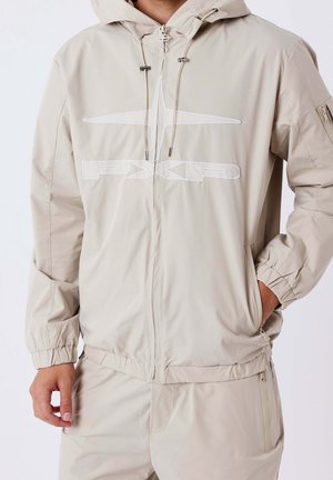 Beige Kapuzenjacke mit Frontreißverschluss, elastischen Bündchen, Kordelzügen, Seitentaschen und PXP-Logo auf der Brust, getragen mit passenden Hosen.