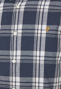 Gros plan sur une chemise à carreaux bleue marine et blanche avec des boutons blancs et un petit "F" brodé en jaune sur la poitrine.