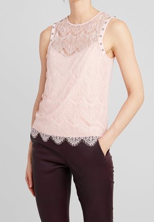 Mouwloze lichtroze kanten top met geschulpte zoom en metalen studs op de schouderbanden, gedragen met donkere strakke broek.