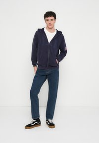 Námořnická mikina na zip s klokaní kapsou, doplněná modrými džíny s přímými nohavicemi a černými teniskami s bílými prvky a gumovou podrážkou.