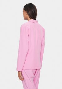 Růžový blazer se strukturovaným designem, s dlouhými rukávy a límcem. Hladká textura a minimální detaily posilují jeho moderní vzhled.