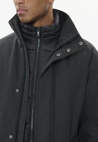 Melna ārējā mētelis ar rāvējslēdzēju priekšā, pogām un augstu apkakli. Iekšpusē ir polsterēts melns jakas ar kviltētu tekstūru.